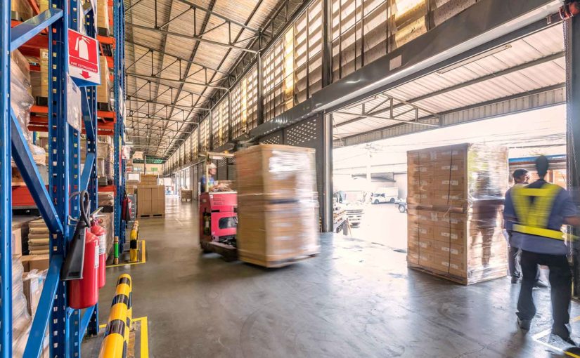 Using a slight motion blur effect, a forklift is shown transporting two large boxes along a loading dock inside a busy warehouse.