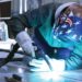 A welding machine operator wearing an American flag helmet welds an aluminum floor on his hands and knees.